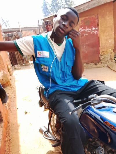 In Toro, Nigeria, a young adult male is seated on a dark blue "BOXER" motorcycle in an unpaved outdoor setting during daylight. He wears a white t-shirt with green trim, a bright blue multi-pocket vest with "Transformed Life Organization" and "Peak" logos, and black cargo pants. White wired earphones are visible, with his left hand positioned near his left ear, appearing to adjust an earbud. His eyes are partially closed, and his mouth is slightly open, suggesting relaxation or drowsiness. Behind him, another male figure wearing a traditional cap is partially visible. The background features a reddish-brown building with a dark red wooden door marked with white text reading "SMAU / NOV / 2014 / 003 / 21," and corrugated metal structures. The ground is light brown dirt.