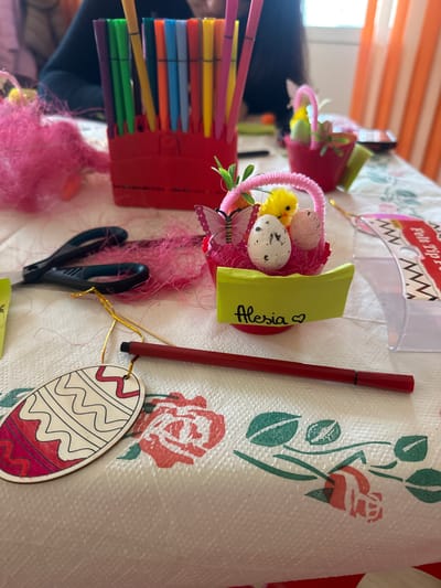 The image shows a tabletop scene with various craft-related objects. There is a container with felt tip pens, small decorative baskets with eggs and chicks, scissors, a marker, and a painted wooden egg ornament. A small name card with "Alessia" is also visible. The tablecloth has a floral pattern with red roses and green leaves. The location is not evident from the image.
