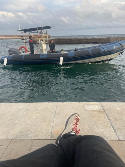 The image depicts a waterside scene in Cabo De Palos, Spain, observed from a low perspective. In the foreground, the lower torso, legs, and right foot of an individual are visible. The individual is wearing black trousers and a light grey shoe with red laces and a white sole, resting on a light-colored, rectangular stone quay or platform.
In the midground, a rigid-hulled inflatable boat (RIB) is afloat on dark green, slightly choppy water. The boat features dark blue inflatable pontoons with a grey stripe, a white hull, and is powered by a Yamaha outboard engine at the stern. A white, metal-framed canopy covers the central steering console. Two individuals are aboard the vessel. One person stands at the helm, dressed in dark attire, possibly a wetsuit, and is positioned behind the steering wheel. The second person is seated further forward in the boat, wearing a dark jacket. A red lifebuoy is attached to the boat's port side, and three white cylindrical fenders hang from the pontoons.
The background reveals a low, rocky breakwater or seawall extending across the horizon, separating the calm water from the open sea. The sky is overcast with scattered clouds.
