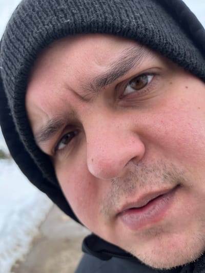 This image, taken in Kalinino, Russia, features a close-up, slightly angled portrait of an adult male. The subject's face is central, displaying fair skin with reddish undertones on the cheeks, brown eyes directed towards the camera, and thick, dark eyebrows that are slightly furrowed. A prominent nose and closed, pale lips are visible, along with light stubble on the upper lip and chin. The individual wears a dark gray knitted beanie hat and the collar of a dark jacket is visible below the chin. The expression is neutral to serious. The out-of-focus background shows patches of white material consistent with snow and darker ground, indicating an outdoor, cold weather environment.