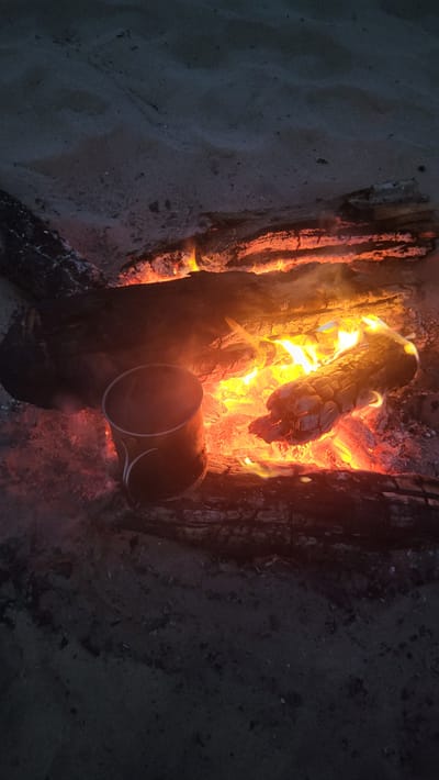 The image depicts a campfire on sandy ground. Several logs are burning, producing an intense orange and yellow glow. A dark metal mug, possibly used for heating a drink, sits near the fire. The surrounding environment is dark and sandy, suggesting an outdoor setting, possibly a beach or campsite. The image appears to have been taken at dusk or night. The location is identified as Zaostrov'e, Russia. There are no people visible in the image.