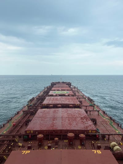 This is a vertical, eye-level, full shot of the deck of a large cargo ship sailing on the ocean under a cloudy sky. The ship's deck is painted a dark red, with multiple large, rectangular cargo hatches stretching towards the horizon. These hatches are also painted red and appear to be wet, with water droplets visible. Yellow markings, including numbers such as "6P," "7P," "6S," and "7S," are painted on the hatches. Pipes and other machinery are visible between the hatches, along with yellow and black striped caution tape. The ocean is a deep blue-green, with moderate waves. In the distance, a few other ships can be seen on the horizon. The sky is overcast with layers of gray clouds. The image conveys a sense of vastness and isolation, with the ship being the dominant subject in a seemingly endless expanse of water and sky. The location is Unknown, Malaysia.