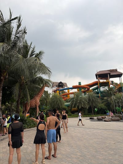 The image depicts a sunny day at a water park, possibly located in Nghĩa Trụ Commune, Vietnam, as suggested by the context. In the foreground, three people are visible from behind. A woman on the left wears a black one-piece swimsuit with a skirt and a light blue visor. Next to her, another woman in a black swimsuit with an open back points to the right. To their right, a man stands in blue swim trunks, with his hands on his hips.
Behind them, a large, decorative giraffe stands amongst palm trees. Further back, a colorful water slide complex with multiple tubes of orange, yellow, green, and blue dominates the background, with thatched-roof structures visible at the top. Several other individuals are scattered throughout the park, some walking and others appearing to be queuing or observing. The ground is paved with interlocking tiles. The sky is overcast with a few hints of blue. The environment appears to be tropical or subtropical, indicated by the presence of lush palm trees and vegetation.