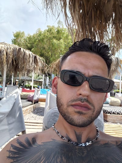 A man wearing sunglasses and a chain necklace is posing for a selfie on a beach. He has a tattoo on his chest. The background shows palm trees and beach loungers. The image is taken outdoors on a sunny day.