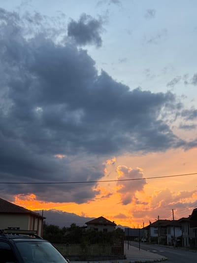 The image captures a dramatic sunset over the city of Blatska, Bulgaria. The sky is a canvas of vibrant orange and yellow hues, pierced by dramatic, dark gray storm clouds. These clouds are edged with a bright golden light, creating a striking contrast. In the distance, a mountain range is visible, its peaks illuminated by the setting sun.
In the foreground, a street runs through the city, lined with houses and trees. The architecture appears to be of a traditional European style, with red-tiled roofs and simple facades. A black SUV is parked in the lower left corner of the frame, partially obscuring a building. Power lines stretch across the scene, adding to the urban feel. The overall atmosphere is one of natural beauty and the transition from day to night, with the storm clouds hinting at impending weather.