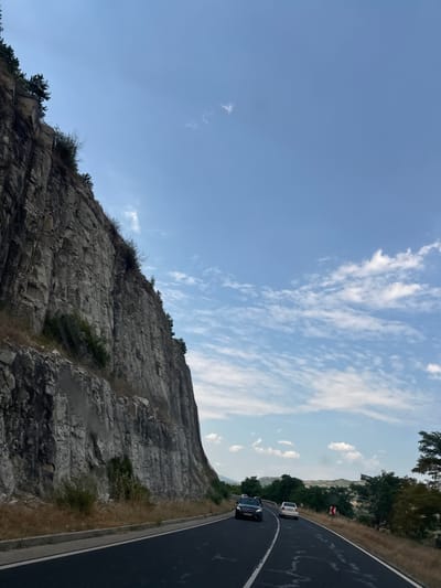 The image depicts a scenic view along a winding road, likely in Zagorsko, Bulgaria. The perspective is from a moving vehicle, looking out towards a dramatic rocky cliff face on the left and the road curving ahead on the right. The sky is a clear, light blue with wispy white clouds scattered across it, suggesting a pleasant, possibly late afternoon or early evening.
On the road, two vehicles are visible: a dark-colored car in the foreground and a white car further ahead. They are traveling along the asphalt road, which has white lane markings. The road is bordered by dry grass and sparse vegetation on the right.
The dominant feature on the left is a massive, layered rock formation, a cliff face that appears weathered and rugged. Small bushes and trees manage to grow in crevices and on ledges of the cliff, adding touches of green to the stone. The rock layers are distinctly horizontal, giving the cliff a stratified appearance.
In the distance, beyond the road and trees on the right, there are hints of rolling hills or mountains, shrouded in a light haze. A small red and white triangular sign is visible by the side of the road, likely indicating a curve or hazard. The overall impression is one of natural beauty and a journey through a mountainous or hilly landscape. There is no visible text in the image.
