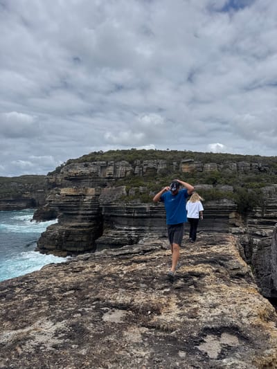 Two people stand on a rocky precipice. One person wears a blue t-shirt and shorts while adjusting something on their head. A second person stands further away, wearing a white top and dark pants. The landscape features layered rock formations and crashing waves below. Sky above is filled with clouds.