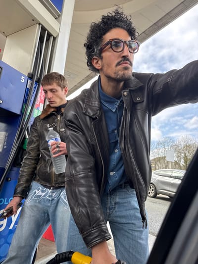 The image captures two males at a gas station in Noisy-le-Sec, France, under an overcast daytime sky. In the foreground, a male with curly dark hair, a beard, mustache, and tortoise-shell glasses, wearing a dark brown leather jacket over a denim shirt and blue jeans, is actively fueling a vehicle, holding a yellow gas nozzle inserted into a partially visible fuel tank. He is looking upwards and to the right, away from the camera. Standing to his left, slightly in the background, is a second male with short light brown hair, wearing a dark brown leather jacket with a fur collar and light blue jeans featuring white graphic designs. He holds a clear plastic water bottle with a blue cap in his left hand, adorned with several rings, and a smartphone-like object in his right hand. He is looking downwards or to his left. Behind him is a blue gas pump dispenser labeled '5', displaying fuel types 'SP98' and 'SP95-E10'. A light-colored SUV is visible in the background beyond the gas station canopy structure.