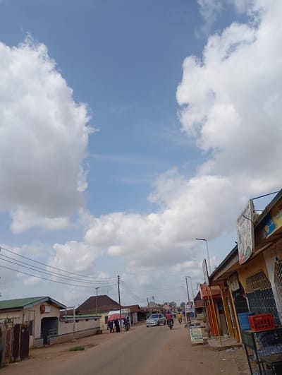 This image depicts a street scene in Kontagora, Nigeria, under a cloudy sky. The street is unpaved and appears to be lined with buildings that are a mix of residential and commercial structures. On the left side of the street, there are low-rise buildings with corrugated metal roofs. One building has a green roof and what appears to be a Coca-Cola advertisement. Further down the street, other buildings with reddish-brown roofs are visible.
On the right side of the street, there are shops with signs, including one that advertises "TANAYUN PROVISION STORE" and "HEALTHY CHOICE." There are also stacks of crates, including a red crate with what appears to be bottles inside, and blue plastic drums.
Several people are present in the scene. Some are walking along the street, and others are on motorcycles. A few motorcycles are seen in motion, and one silver car is driving down the street. Two individuals are standing under a red umbrella. Overhead, power lines are strung across the street, supported by utility poles. The overall impression is that of a bustling, typical street in a Nigerian town.