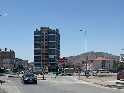 In Kayseri, Turkey, a street scene unfolds under a clear blue sky. A tall, modern building, labeled "UZUM PLAZA," dominates the mid-ground, its grey facade contrasted with bands of brown and numerous windows. Across the street, a sign for "ERCIYES HASTANESİ" (Erciyes Hospital) is visible, with smaller text below reading "ACİL" (Emergency). A black Audi sedan is driving through an intersection in the foreground, its headlights on. To the left, two buses are stopped at what appears to be a bus station. A lone pedestrian is crossing a crosswalk between the buses and the hospital sign. In the background, residential buildings and a large, arid mountain range suggest the city's geographical setting. Traffic signs, including "DUR" (Stop) signs, are present. The environment appears to be an urban commercial and residential area.