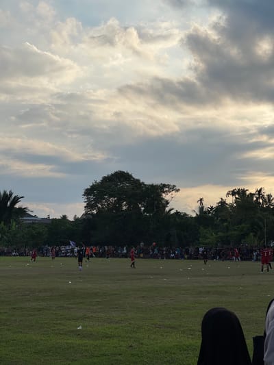 The image depicts an outdoor soccer match taking place in Aratontonmoncrang, Indonesia. A large crowd of spectators lines the field, some sitting and others standing. Several players are visible on the green grass field, some in red and white uniforms, others in black. The sky above is filled with dramatic clouds, with hints of sunlight breaking through, suggesting either early morning or late afternoon. Lush green trees form a dense backdrop to the scene, with palm trees and other foliage visible along the perimeter of the field. Some white debris is scattered across the grass. In the foreground, out of focus, are parts of spectators.
