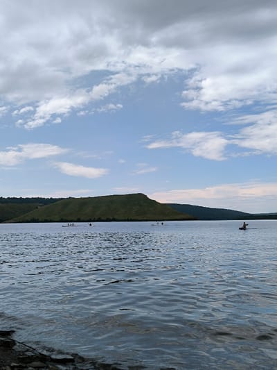 The image displays a wide view of a large body of water, likely the Dniester Reservoir, near the city of Horaivka, Ukraine. The foreground consists of dark, rippled water, with a partial view of a rocky shoreline at the bottom left. In the mid-ground, a substantial, verdant green hill with a rounded summit occupies the central and left horizon, receding into additional darker hills on the right. The sky above is a mix of light blue and various cloud formations, including scattered white cumulus clouds and larger, more diffuse grey clouds towards the upper edges. On the water's surface, several recreational vessels are visible. In the mid-left distance, a cluster of approximately three light-colored kayaks or canoes with individuals aboard are present. To the mid-right, a single individual in a darker-colored kayak is observed paddling.