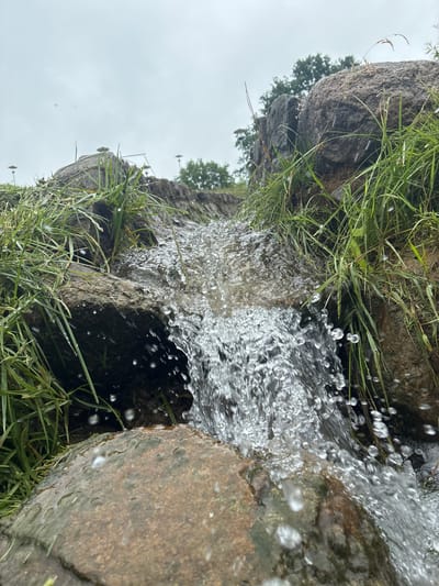 The image shows a close-up of a small waterfall flowing over rocks and grass. The water is clear and appears to be in motion, with droplets visible in the air. Green grass and brown rocks surround the waterfall. The sky is overcast and appears to be in the background. The location is identified as Sabile, Latvia. There are no people or animals visible in the image.