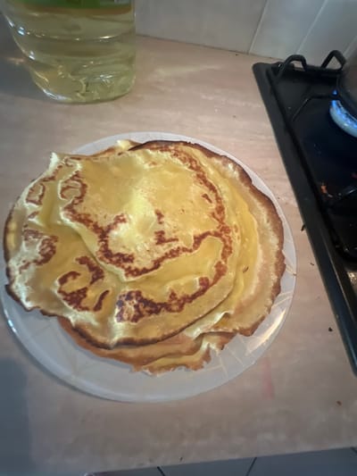 A close-up shot shows a stack of freshly cooked crepes on a white plate. The crepes are golden yellow with slightly browned edges, and their surfaces show the characteristic irregular patterns of this dish. A bottle of cooking oil is visible in the upper left corner, and the edge of a gas stovetop with a blue flame is in the upper right corner. The scene appears to be set on a kitchen countertop, suggesting a home environment. The image is well-lit, highlighting the textures of the crepes. The location of the image is not specified in the provided information.
