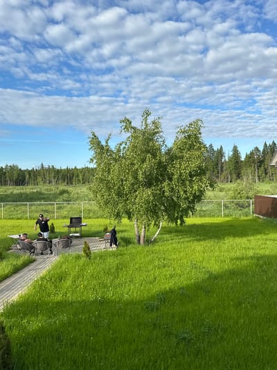 The image displays a vibrant outdoor scene under a bright blue sky filled with numerous scattered white clouds. A wide expanse of lush green grass dominates the foreground and midground, indicative of a well-kept lawn. In the center, a prominent birch tree with abundant green foliage stands tall, casting a light shadow on the grass.
In the background, a dense line of tall, dark evergreen trees forms a natural border, suggesting a forest. Above the treeline, a small building with a dark brown roof is partially visible. A white fence runs horizontally across the midground, separating the manicured lawn from a patch of wilder vegetation that leads to the forest.
A paved pathway, made of light-colored pavers, curves through the grassy area. Along this path, several outdoor amenities are arranged for a gathering: a barbecue grill, a table, and multiple chairs, some of which are reclined. A small wooden bench, with a dark jacket draped over it, is also present.
At least three individuals are visible on the pathway. One person, wearing a black t-shirt and a cap, stands near the chairs, while at least two others are seated in the reclining chairs. The arrangement of the grill and seating strongly suggests a casual outdoor event, such as a barbecue or a relaxed social gathering.
The bright sunlight and clear sky indicate it is daytime, likely late morning or afternoon, perfect for outdoor activities. The overall atmosphere appears tranquil and leisurely. No text is visible in the image. The setting, with its expansive lawn, forest backdrop, and birch tree, is characteristic of a rural or semi-rural environment, aligning with the given location context of Веревское, Russia.