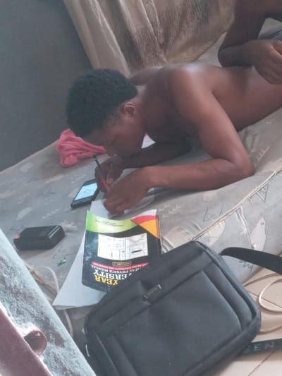 Here's a concise description of the scene:
A shirtless person lies on a mattress, head supported by a pink cloth, while holding a pen and phone, seemingly engaged in writing or studying near a book titled "Year University Physics Manual". A black bag is situated nearby, on what appears to be the mattress. The setting is a room with patterned bedding or a mattress with a patterned cover. The location Achara Ndiagu Omegio, Nigeria, cannot be verfied in the image.