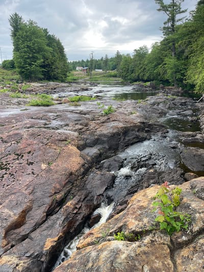 The image depicts a rocky riverbed with a stream flowing over the rocks. The river is surrounded by lush green trees and a cloudy sky. In the distance, there are buildings that appear to be part of a rural settlement. The foreground is dominated by large, weathered rocks with patches of moss and small plants growing in crevices. The water is clear and shallow in some areas, with small rapids and waterfalls forming as it navigates the rocky terrain. The overall scene conveys a sense of natural tranquility and a remote, possibly wooded, location.