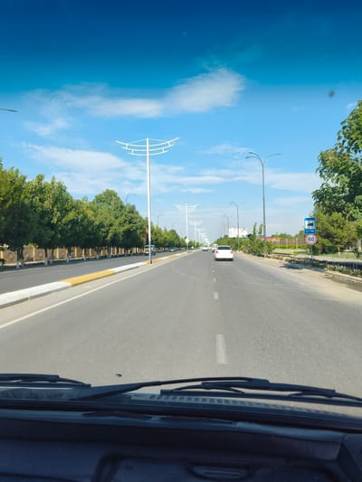 This media file captures a view from inside a vehicle driving down a paved road lined with trees in Buz, Uzbekistan. The road has clear lane markings and a yellow and white striped curb on the left. Tall, modern streetlights with curved tops are visible along the road, suggesting an urban or suburban setting. On the right side of the road, there are more trees and some signage. One sign clearly displays "RADAR" and a circular sign with "60", indicating a speed limit of 60.
Several vehicles are on the road, including a white sedan ahead and another vehicle on the left. The scene appears to be set during the daytime, with a bright blue sky and scattered white clouds, suggesting fair weather. The trees are green and leafy, indicating a warm season. The overall impression is of a routine drive through a well-maintained road infrastructure in a location that appears to be well-developed. There are no people visible within the frame of the road and surrounding environment. The emotion conveyed is one of normalcy and travel.