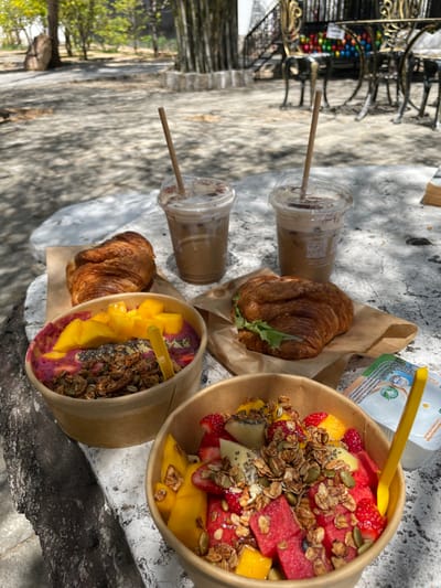 The image displays an outdoor dining scene featuring a breakfast or brunch spread laid out on a light-colored, mottled table, possibly made of stone or concrete. The setting appears to be outdoors, likely in a cafe, park, or garden area, characterized by dappled sunlight and shadows from overhead foliage.
In the foreground, from left to right across the table (mentally reorienting the image to a standard horizontal perspective), there are several items:
Two clear plastic cups with iced beverages, likely iced coffees or lattes, complete with paper straws. One of the cups has a QR code visible on its side.
Next to the drinks, there are two croissants. One is a plain, golden-brown pastry. The other is a croissant sandwich, with green lettuce peeking out from the middle, suggesting a savory filling. Both croissants are resting on pieces of brown paper.
Further to the right, two colorful bowls are presented, made of brown paper or cardboard.
The bowl on the right is filled with a vibrant fruit salad, prominently featuring cubes of red watermelon, yellow mango chunks, sliced red strawberries, and slices of green kiwi. It is generously topped with a mix of granola, pumpkin seeds, and chia seeds. A yellow plastic spoon is inserted into the bowl.
The bowl on the left contains what appears to be a smoothie bowl, with a deep purple base, topped with bright yellow mango pieces, and a similar granola/seed mixture. This bowl also has a yellow plastic spoon.
Behind the fruit bowl, a small, flat-topped white container with a yellow lid and a yellow plastic spoon is visible. The label on this container features a green leaf logo and clear text indicating "VEGAN" and "BIO," along with other illegible text.
The table surface itself is bright, with prominent shadows of leaves suggesting direct sunlight filtering through trees. In the blurred background, there are hints of decorative dark metalwork, possibly part of chairs or railings, and further indistinct greenery and sunlit ground, reinforcing the outdoor, casual atmosphere. The overall impression is one of a fresh, healthy, and pleasant outdoor meal on a sunny day.