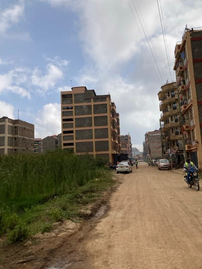 A vertical shot depicts a dusty road flanked by developing buildings under a partly cloudy sky. The buildings, a mix of completed and under-construction structures, line both sides of the road. Vehicles, including cars and motorcycles, are parked or moving along the road. A grassy area borders the road on the right side, with a dirt bank separating it from the road surface. Utility lines stretch across the sky above.