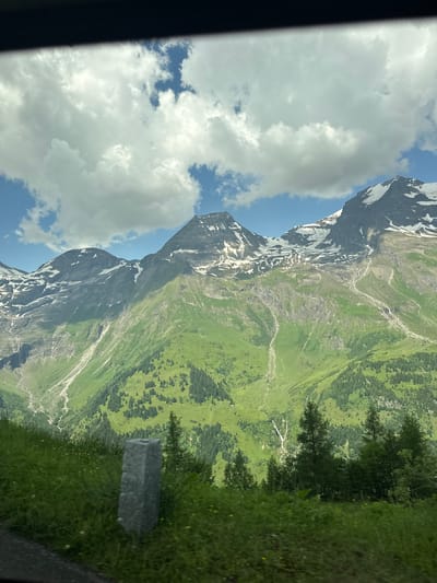 The image shows a mountainous landscape in Fusch an der Großglocknerstraße, Austria. The mountains are covered in green grass and scattered trees, with patches of snow on the higher peaks. The sky is blue with fluffy white clouds. In the foreground, there is a stone marker and some tall grass.