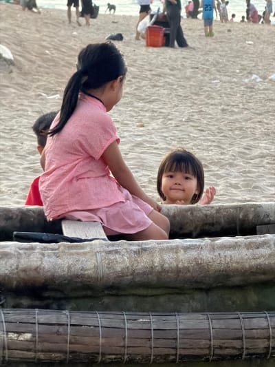 Two young girls are positioned near a structure composed of weathered, cylindrical posts, possibly part of a building or boardwalk in Thuận An Ward, Vietnam. One girl, with dark hair in a ponytail and wearing a striped pink shirt, is seated while reaching out. The other girl, with short dark hair, is peeking out from behind the structure, her expression inquisitive. In the background, a sandy beach stretches out with blurred figures of people scattered across the landscape. The overall environment suggests a sunny day at the beach.