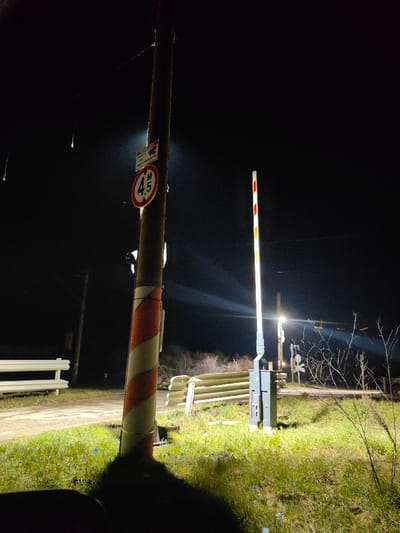 This image depicts a railway crossing at night in the city of Dralfa, Bulgaria. The scene is illuminated by artificial light sources, casting distinct beams and reflections. In the foreground, a prominent utility pole, marked with red and white diagonal stripes near its base, stands on the left side of the frame. Attached to the pole are two signs: a circular white sign with a red border indicating a 4.5-meter height restriction ("4.5 M"), and a smaller rectangular sign above it displaying a camera icon with Bulgarian text "ОБЕКТЪТ Е ПОД ВИДЕОНАБЛЮДЕНИЕ" (indicating video surveillance).
To the right of the utility pole, a railway crossing barrier arm is positioned upright, signifying an open crossing. This barrier arm is illuminated and features reflective red and white stripes. Its grey control mechanism housing is visible at its base. Behind the barrier, white corrugated metal guardrails are visible, running parallel to what appears to be a dirt or gravel path. Further in the background, more railway signs, including a crossbuck, are discernible under the glare of distant lights. The ground in the foreground and around the crossing consists of green grass and some dry, sparse brush. The sky is dark and clear, typical of nighttime conditions. No individuals or active vehicles are present in the frame. A dark, undefined mass occupies the bottom left corner, suggesting a part of the immediate foreground or possibly an obscured vehicle interior from which the photo was taken.