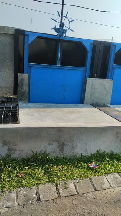 The image depicts a blue sluice gate structure, likely part of a water management system, in a location that appears to be the city of Sidodadi, Indonesia. The gate is open, revealing a dark interior. Above the gate, a blue steering wheel mechanism is visible, used for operating the gate. The structure is made of concrete and metal, with some of the concrete showing signs of weathering and staining. In the foreground, a small patch of grass is growing along a concrete border, with some debris, including a wrapper, scattered on the grass. The ground is a mixture of concrete and gravel. Overhead power lines are visible against a clear, light-colored sky. To the left of the gate, a metal grate is positioned on the concrete. The overall scene suggests an outdoor environment, possibly a canal or drainage area, in an urban or semi-urban setting.