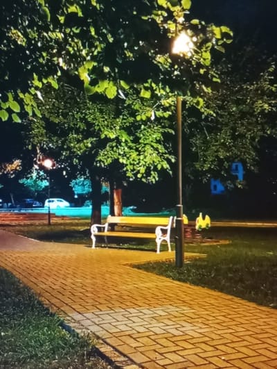 This nighttime image depicts a public park or walkway in Mirbozor, Uzbekistan. A brick-paved walkway, laid in a herringbone pattern, extends from the lower left foreground, illuminated by artificial light. Green grass borders both sides of the pathway. To the right of the walkway stands a black lamppost with a glowing light source, casting illumination on the immediate area. Adjacent to the lamppost and to the left of the path, an empty park bench with a yellow slatted seat and white ornate metal armrests and back supports is positioned. Behind the bench and lamppost are dense trees with green foliage, prominently lit. Further to the right, on a grassy patch, a yellow spring rider is partially visible. In the background, beyond the trees, a faint white vehicle and other indistinct structures or buildings are discernible, along with distant streetlights. The sky is dark, indicating night. No individuals are present, and no actions or interactions are occurring within the visible scene.