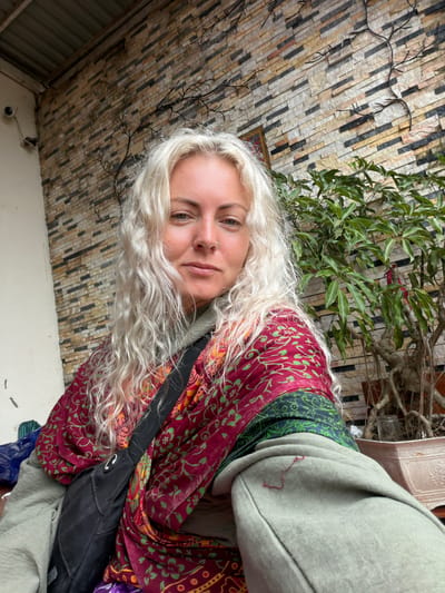 A young Caucasian female with long, wavy blonde hair is seated outdoors. She is wearing a light green hoodie with a decorative red and green patterned scarf draped over her shoulders. A black crossbody bag is positioned in front of her. She has a nose ring and is looking directly at the camera. The background features a wall constructed of textured, multi-colored rectangular stones, with dark, branch-like decorative elements on its surface. To the right, a lush green plant in a pot is visible. The scene appears to be in Hướng Phùng Commune, Vietnam.