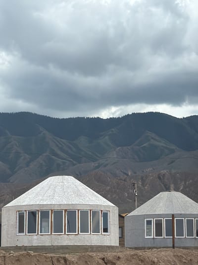 The image depicts a landscape scene with two yurt-like structures in the foreground, set against a backdrop of rolling, sparsely vegetated hills and mountains under a cloudy sky. The structures appear to be under construction or recently completed, with rough surfaces and visible windows. The leftmost structure is a rounded building with a conical roof, featuring a series of rectangular windows along its side. To its right, a second similar structure is visible, also with a conical roof and windows. Between the two structures, a utility pole is present. The ground in the foreground is dry and appears to be earth or sand. The overall environment suggests a semi-arid or mountainous region. The context of Kaji-Say, Kyrgyzstan, implies a location in Central Asia known for its mountainous terrain and traditional dwelling styles.