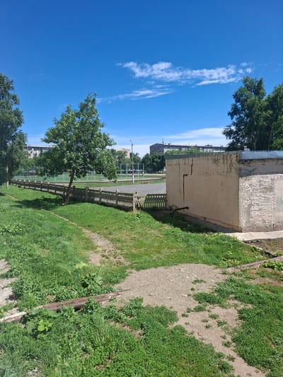 The image shows an outdoor scene with a bright blue sky and scattered white clouds. In the foreground, there is a patch of overgrown green grass with a dirt path winding through it. A rusty pipe lies partially embedded in the grass. To the right of the grass, there is a pale, weathered concrete building with a cracked wall. A decorative concrete fence runs horizontally across the middle ground, separating the grassy area from a sports field. The sports field, which appears to be a basketball or volleyball court with blue markings, is visible beyond the fence. In the background, several multi-story apartment buildings and trees are visible under the expansive sky.