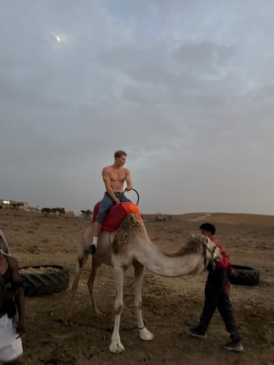 A young, shirtless, muscular man with blond hair is seated atop a camel, holding onto a black reins-like object. He is wearing blue denim shorts and white socks. The camel is light brown and white, with a red saddle pad. To the right of the camel, a person wearing a red shirt and dark pants is standing and appears to be interacting with the camel. In the background, there is a dry, dusty, hilly landscape with sparse vegetation and some distant buildings. The sky is overcast with a sliver of moon visible. The ground is covered in dry grass and dirt, with a large tire visible in the lower left corner. The scene suggests a rural or desert environment, possibly near Douar Ait Daoud, Morocco.