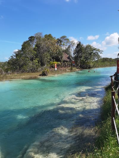 The image shows a view of a turquoise-colored lagoon in Unidad de Riego, Mexico, located at latitude 18.591 and longitude -88.434. The lagoon is bordered by lush vegetation and a sandy bank on the right, with a wooden fence running parallel to the water’s edge. Two individuals are swimming in the center of the lagoon. A small thatched hut is visible in the background on the left, near a small, grassy clearing and a dense stand of trees. The sky is clear blue, and the sun is shining. The scene is calm and peaceful. The vegetation is green and lush. The water is clear and inviting, and the sky is blue and cloudless. The overall impression is one of tranquility and natural beauty.