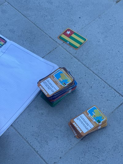 The image displays a close-up, aerial view of a group of educational cards spread across a grey tiled surface, possibly a patio or walkway. The tiles have a speckled texture and are laid in a grid pattern. To the left of the frame, a portion of a spiral-bound notebook with graph paper is visible, partially covering the tiles.

Dominating the scene are stacks of colorful cards, each featuring a map of a country and text in a foreign language. One card, partially obscured, shows a map of Africa and the word "АФРИКА". Another card, more clearly visible, displays a map of Madagascar with text in Ukrainian, including "Мадагаскар" and "Перт-Луї", and statistical information. Further to the right, a stack of similar cards is visible, with one card prominently showing a map of Zambia and the city "Лусака". The text on this card is also in Ukrainian, with details about the country's area and population.

In the upper right quadrant of the image, a single card depicting the flag of Togo is laid out flat. The flag has horizontal stripes of green and yellow, with a red square in the upper left corner containing a white star.

There are no people visible in the image. The lighting suggests it is daytime, with some natural light illuminating the scene. The overall impression is of an educational activity or game taking place outdoors.

The location of Yablunivka, Ukraine, is not discernible from the visual information presented in the image.