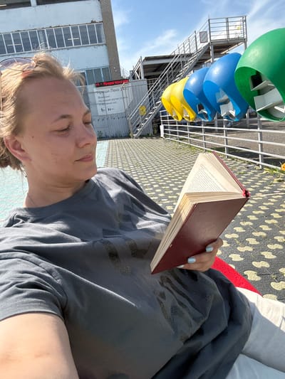 A woman is lying down, reading a book, outdoors in what appears to be the city of Sin’kovo, Russia. The book has a red cover and open pages. The woman has blonde hair and is wearing a gray T-shirt. The location is a public space; to the left is a multi-colored sculpture of what appear to be green, blue, and yellow funnels. There is a building with a sign containing Russian text that reads "Kryshi Leko 24x7". The ground is patterned.