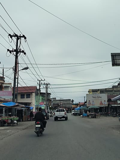 The image captures a street scene in the city of Simpangpeut, Indonesia, under an overcast sky. A power pole with numerous wires dominates the left side of the frame, with a street lamp attached. Several buildings line the street, some featuring shop fronts with signage. One building displays a pink sign with the text "ftfren" and "CELL". Further down the street, a large billboard with the text "Idul Adha" is visible, featuring images of four people.

Several vehicles are present on the road. A man is riding a motorcycle with a license plate that reads "BL 3618 HAP". A white pickup truck is also on the road, and a white van is parked. To the left of the road, there are market stalls with blue and green tarps, displaying various goods. A few people are standing near the stalls and on the sidewalk. The road surface is asphalt, and the ground beside the road is a mixture of dirt and debris. The overall environment appears to be a commercial area of the city.