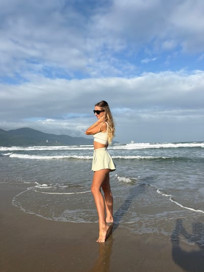 The image displays a full-body view of an adult female standing barefoot on a sandy beach within An Hải Ward, Vietnam. She is positioned at the water's edge, with foamy white waves breaking around her feet. The subject has long, wavy blonde hair and wears dark sunglasses, a light-colored sleeveless crop top, and a light green pleated mini-skirt. Her right arm is raised, with her hand near her left shoulder, while her left arm is positioned behind her back. She is turned slightly, looking over her right shoulder away from the camera. The background consists of the ocean with moderate waves, extending towards distant green mountains under a partially cloudy blue sky. A small white object, possibly a distant sailboat, is visible on the water to the far left, and a larger white vessel is present further out to sea on the right.
