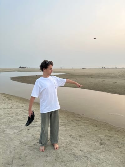 An adult male stands barefoot on a sandy beach in Betalbatim, India, facing right and pointing into the distance with his right arm extended. He possesses curly brown hair, with dark sunglasses resting on top of his head. He wears a white short-sleeved t-shirt with a small dark graphic on the left chest and loose-fitting olive-green or grey-green wide-leg pants. In his left hand, he holds a single black shoe.

The beach environment features a shallow, winding water body, possibly a stream or tidal pool, running from the mid-left towards the mid-right foreground. Footprints and tire tracks are visible in the sand around the individual. In the distant background, the sandy shoreline extends under a hazy, pale sky, where several small groups of people are dispersed. Two fishing boats are partially visible on the far left horizon. A dark, kite-like object or bird is in flight in the upper right quadrant of the sky.