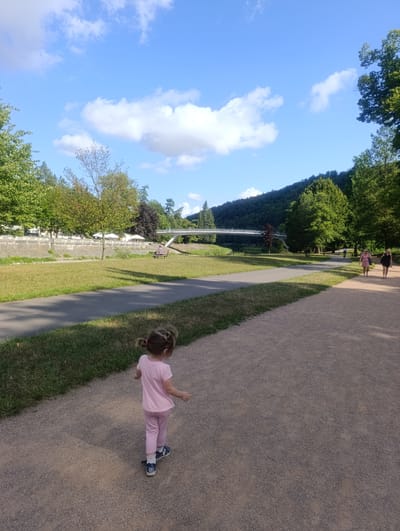 A young girl with blonde hair in pigtails, wearing a pink t-shirt and pink pants, walks away from the camera on a gravel path. She is wearing white socks and blue shoes with the number "01" on them. The path curves to the right and runs alongside a grassy area. In the background, there is a river, a footbridge, a hill covered with trees, and a building with a row of white umbrellas in front of it. Two women are walking on the path in the background, and a person is sitting on a bench in the distance. The sky is blue with scattered white clouds. This scene appears to be in Teplice nad Bečvou, Czechia, a location known for its spa and parkland.