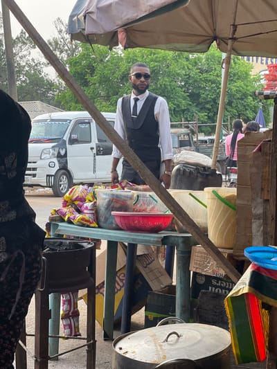 The image captures an outdoor market or street vending scene during what appears to be bright daylight. The perspective is from a low angle, looking towards a vendor and his stall.
In the right half of the image, a man stands attending to his goods. He is dressed in a white long-sleeved shirt, a dark vest, and a dark tie, suggesting a somewhat formal appearance for a street vendor. He has a beard and is wearing dark sunglasses, obscuring his eyes. His right hand is visible, resting on or near items on his stall.
The stall itself is a rustic setup, constructed from metal poles and wooden planks. It holds various items for sale. On the left side of the stall (from the viewer's perspective), there are transparent plastic containers; one appears to be filled with light-colored granular material, possibly rice or other grains, while another has a red lid. Below these, multiple plastic bags filled with snacks or chips, featuring yellow and purple packaging, are displayed. On the right side of the stall, a large metal pot with a lid is visible. Above it, a blue plastic basin and a piece of striped fabric (with green, yellow, and red colors) are placed. Several cardboard boxes are stacked behind the main display, with partial text visible, including "Lion Butters," "RITE," and "Dais."
In the background, behind the vendor and his stall, green trees indicate an outdoor setting. Several vehicles are parked or moving; a white van or mini-bus is prominently visible. Other people are faintly discernible in the upper left background, including one person with long, braided hair.
In the immediate foreground, at the bottom of the image, the back of a person wearing a dark, patterned garment is visible, suggesting the photograph was taken from behind another individual who might be seated or observing. The overall atmosphere suggests a typical bustling market environment.