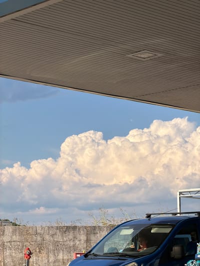 An eye-level, wide shot captures a sky filled with voluminous white clouds under a bright blue sky. A large, ribbed canopy juts into the frame from the top, casting a shadow over the scene. Below the clouds, a weathered concrete wall runs horizontally across the middle ground. To the left, a red fire hydrant stands beside the wall. In the foreground, the hood of a dark blue van is visible, with a person holding a phone inside the driver's seat. The van is positioned in front of the wall and the fire hydrant. Tall, dry grasses sprout from behind the wall. In the far background, a white structure with horizontal beams can be seen. The overall lighting suggests it is daytime.