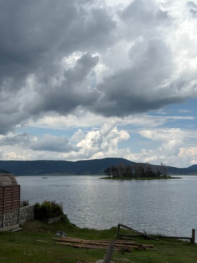 This wide-angle landscape photograph depicts a scene at the Batak Reservoir, located near the town of Цигов чарк, Bulgaria, under a dynamic sky. The upper two-thirds of the image are dominated by a heavy, turbulent cloud cover, transitioning from dark grey, dense cumulus clouds in the upper left to brighter white clouds and intermittent patches of blue sky towards the upper right and central horizon.
The mid-ground features a large body of water, its surface exhibiting numerous small ripples, reflecting the subdued light from the sky. Approximately one-third across from the left edge and halfway down from the top, a small, light-colored boat carrying three to four indistinct human figures navigates the water. Towards the right side of the water body, in the mid-ground, a small, irregularly shaped island is present, densely covered with dark, deciduous trees, some appearing sparse or bare-branched. Beyond the water, a continuous line of dark, forested hills or mountains extends across the horizon, forming the distant background.
In the immediate foreground on the left, a partial view of a building is visible. It features a facade constructed with horizontal wooden slats above a stone-stacked wall base. Adjacent to the building, a small area of green grass slopes gently towards the water. On this grassy incline, a pile of approximately ten cut logs of varying lengths is laid out horizontally. To the right of the logs, a rustic wooden fence, appearing partially broken or unfinished, extends towards the water's edge. A dark, possibly bundled object, resembling a tarp or folded fabric, lies on the grass near the logs. Green bushes and other foliage are present between the building and the water, partially obscuring the immediate shoreline. No clear human activity or interaction is depicted beyond the distant boat.
