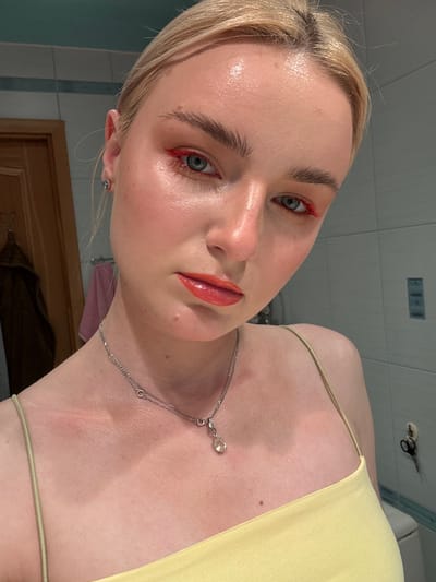A young Caucasian woman with blonde hair pulled back is shown in a selfie. She is wearing a pale yellow tank top and a silver necklace with a clear pendant. Her makeup includes bright red eyeliner and orange lipstick. She is facing the camera directly, with her head slightly tilted. The background appears to be a bathroom with tiled walls and a wooden door. There is a hint of a light fixture in the upper right corner. There is no indication of the location in Sinyavino, Russia.