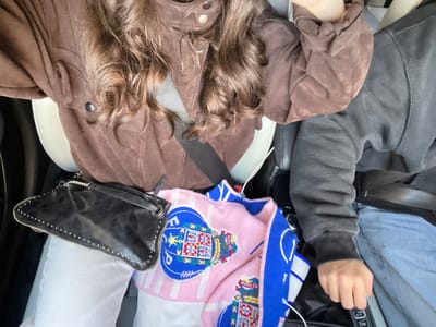 Two people are in a car. The passenger is wearing a brown suede jacket, with curly hair visible. A black studded purse rests on their seat along with a folded pink and blue patterned knit. The driver, wearing jeans and a gray hoodie, grips the gear shift. The seats are white.
