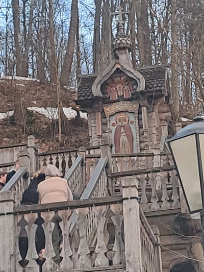 A tilted medium shot shows a wooden structure next to a leafless tree in Vzglyadnevo, Russia. The structure features intricate carvings and a painting of a figure with a halo, possibly a religious icon. A partial view of a person in dark clothing and a light-colored top is visible on the structure. A lamppost sits atop the structure. The environment is mostly natural with bare trees and a hint of sky in the background.