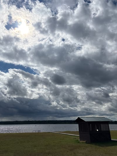 The image captures a scenic view of a lake under a dramatic sky with a mix of sun and dark clouds. In the foreground, a small, dark wooden structure with a corrugated metal roof stands on a grassy area. A wooden walkway leads from the structure towards the lake. The lake's surface is rippled and reflects the sunlight, with a distant treeline forming the horizon. A few distant figures are visible on the water, possibly paddleboarders. The overall impression is a natural landscape with a man-made element, suggesting a park or recreational area near a body of water. The sky is filled with large, billowing clouds, some of which are dark and foreboding, while others are illuminated by the sun breaking through.
