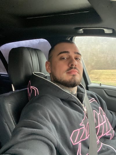 A male individual is depicted seated in the driver's compartment of a vehicle, located in Mikre, Bulgaria. The subject possesses short dark hair, a beard, and visible tattoos on his neck and left temple. He wears a black zippered hoodie with prominent pink embroidered designs on the front and a smaller corresponding design on the upper left shoulder. A diagonal seatbelt secures him across the torso. The vehicle's interior features black leather seating and a dark headliner with an integrated sunroof. The subject is facing forward, looking directly at the camera. Through the passenger side window, an indistinct outdoor environment is visible, characterized by distant trees and open ground under an overcast sky.