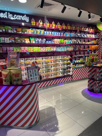 The image showcases a candy store display. The scene is lit with spotlights on the ceiling. A purple wall serves as the backdrop for the shelves holding candy. A neon sign reading "red is.candy" is located on the left side. The shelves are stocked with various candies including Haribo, and other packaged sweets. In the foreground, there are bulk candies in clear containers, along with bagged candies. A sign advertises "Granel $14.990" for 500 grams. Striped red and white decorations are on the lower and right side of the image. The location is implied to be in Pudahuel, Chile, based on the OCR.