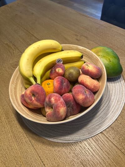 A fruit bowl filled with bananas, peaches, tangerines, kiwis, and a mango sits on a wooden table. The fruit bowl is made of bamboo and rests on a woven placemat. The table has a light brown, wood-grain finish. In the background, a dark chair is partially visible. This scene is located in Oldemarkt, Netherlands.